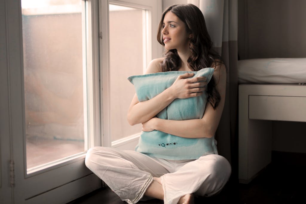 Serene woman sitting indoors, embracing pillow, lost in thought by a bright window.